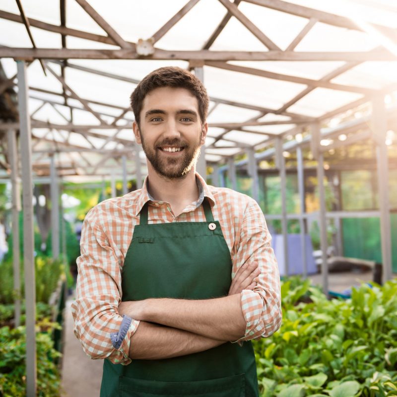 Young handsome gardener smiling, posing with crossed arms among flowers.